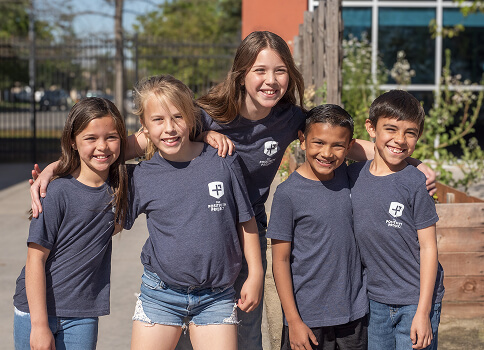 5 children wearing Positivity Project shirts smiling outside of a school
