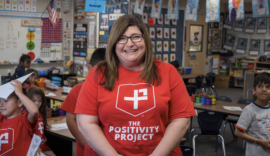 A smiling woman in a classroom wearing a Positivity Project shirt, surrounded by students