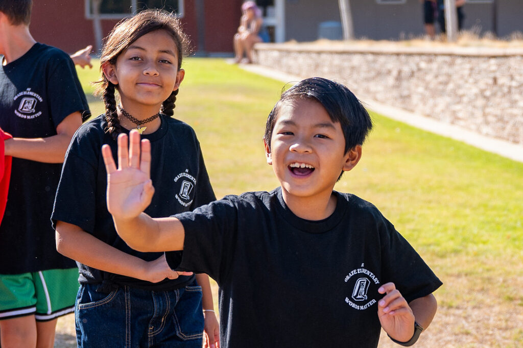 one-student-waves-at-camera-with-another-student-in-background